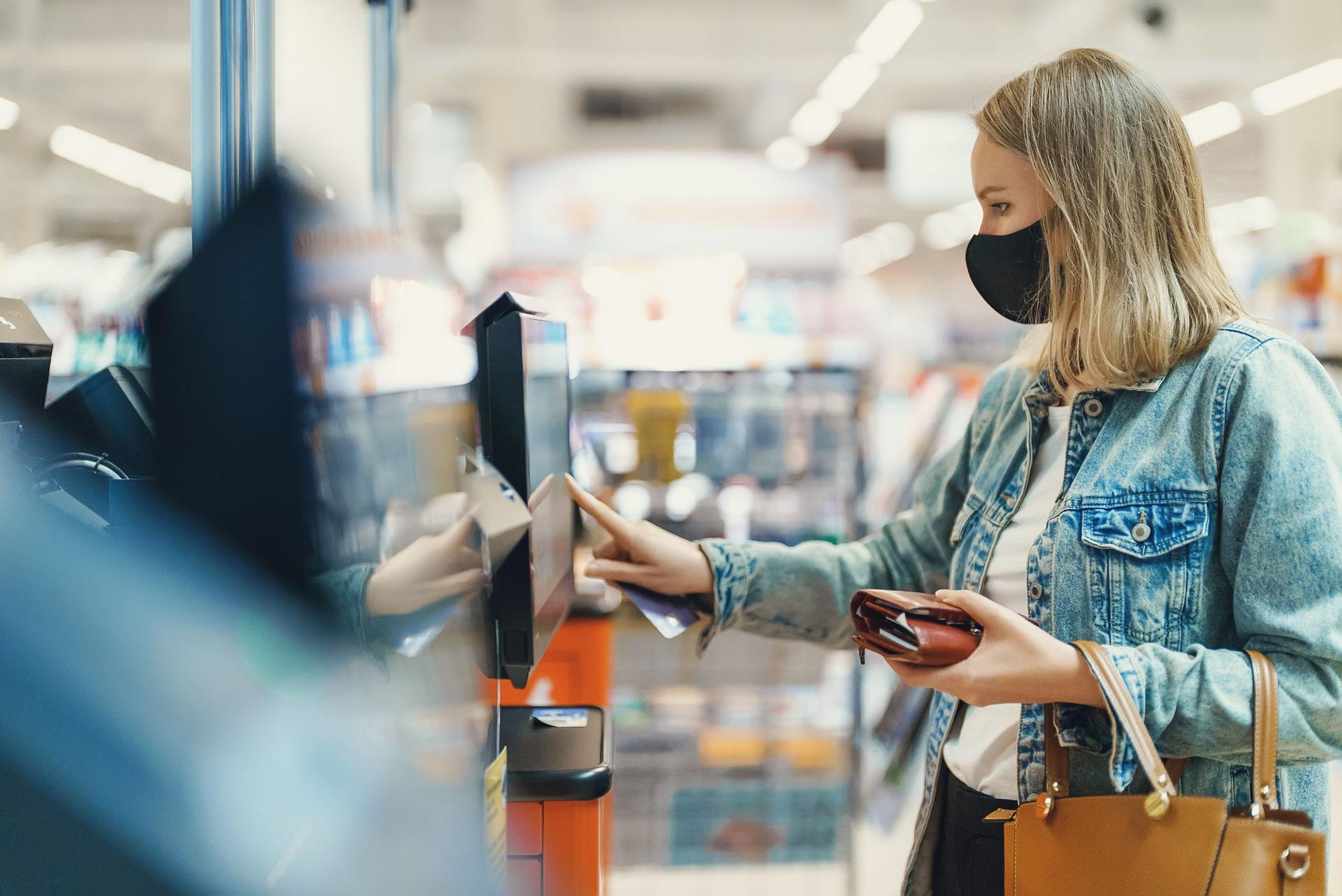 Woman in medical mask pays at self-checkouts.